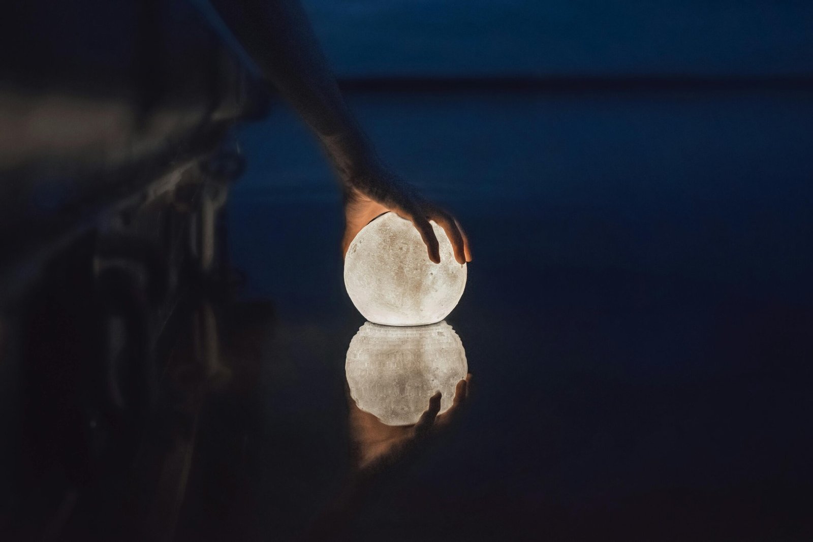 A hand gently touches a glowing sphere resembling the moon, reflecting on dark water.