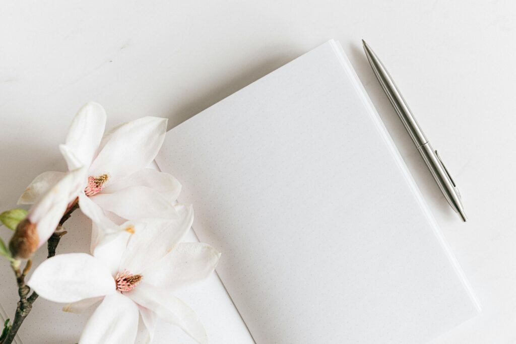 Top view composition of opened notebook with blank white sheets and stylish silver pen decorated with lush blooming Magnolia twig placed on white background
